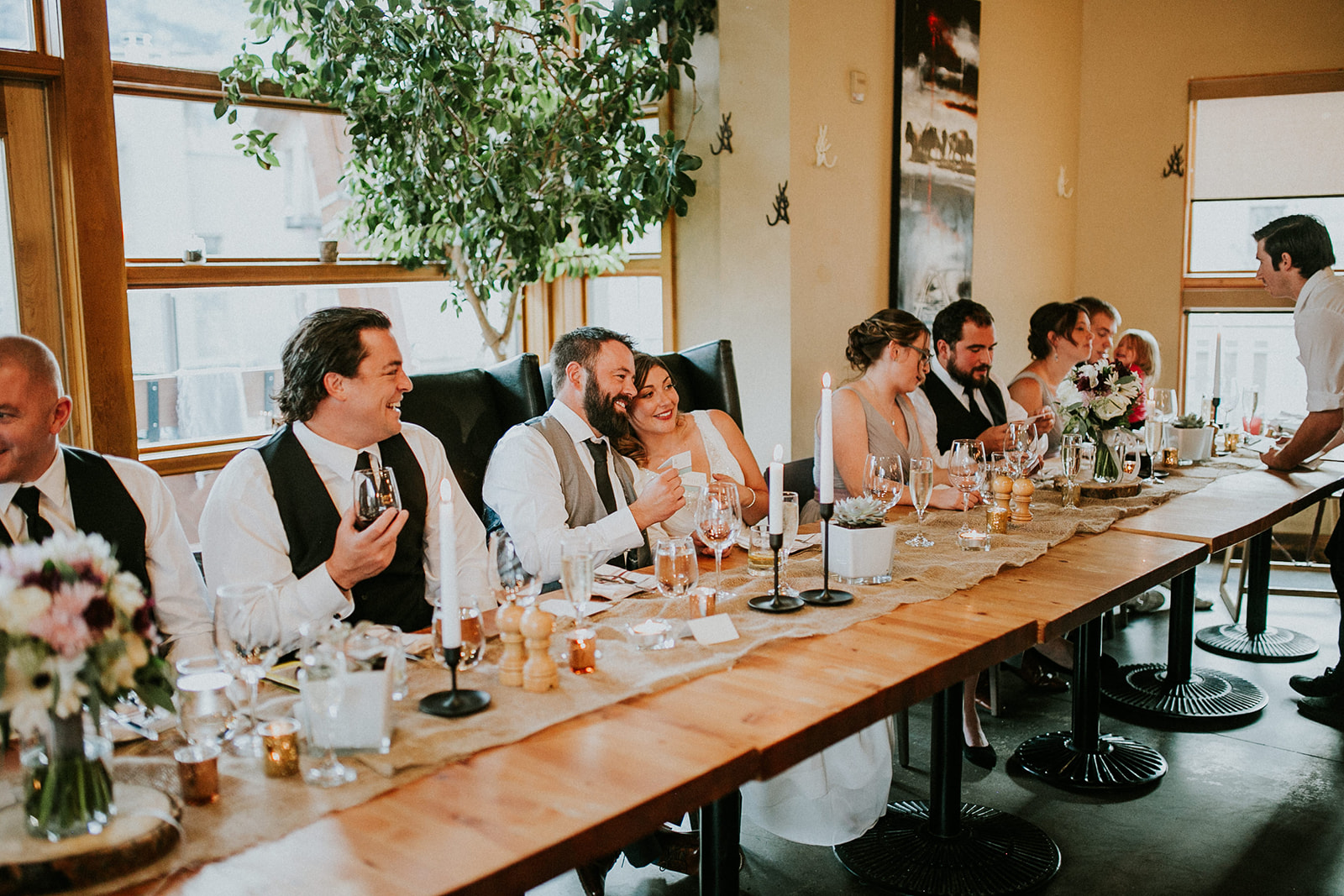 A wedding party sits at a long wooden table, laughing and smiling together. The bride and groom are seated in the center, surrounded by their guests, as a photographer captures the joyful moments among flowers, candles, and glasses.