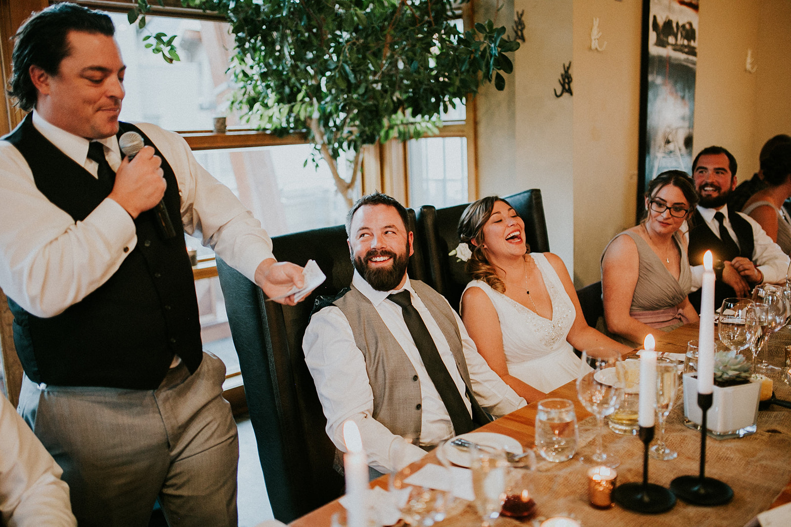 A photographer captures a man in a vest giving a speech with a microphone at a Calgary wedding reception. The bride and groom, seated at a candlelit table, laugh and smile along with their bridal party.
