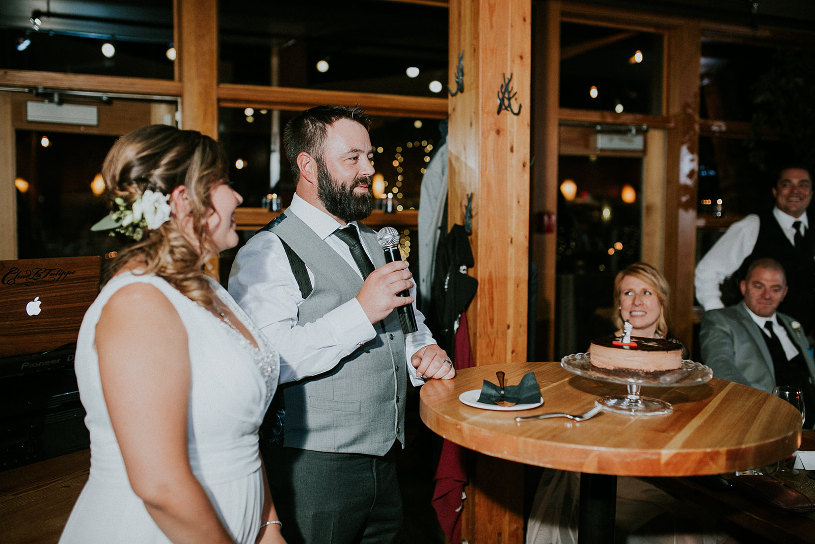 A man in a vest and tie speaks into a microphone at a wooden table with a cake, while a woman in a white dress stands beside him and guests listen—captured by the photographer in a warmly lit indoor setting in Banff.