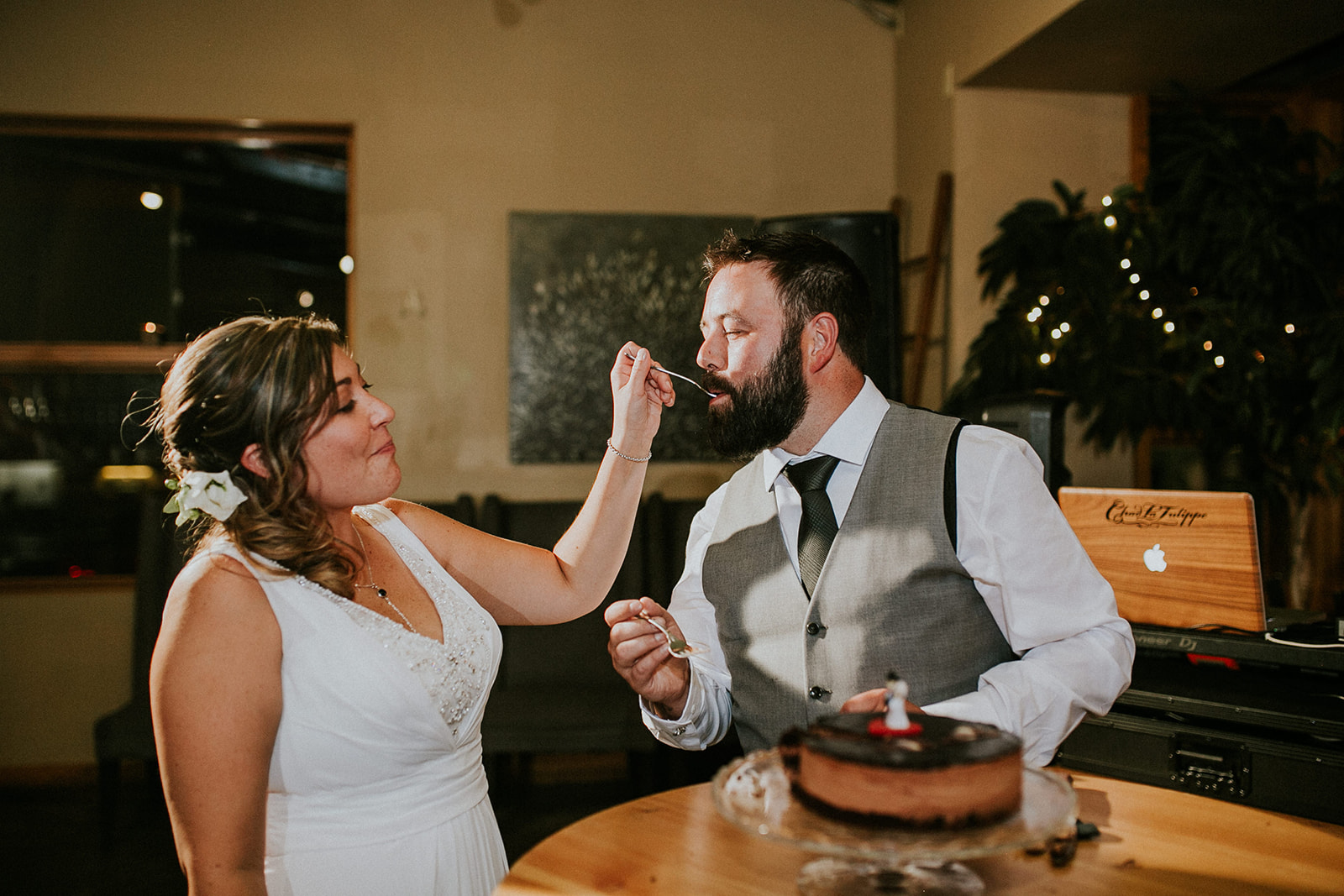 A bride in a white dress feeds cake to a smiling groom at their wedding reception. A chocolate cake sits on the table, with decorations, a laptop, and their Canmore photographer capturing every joyful moment.
