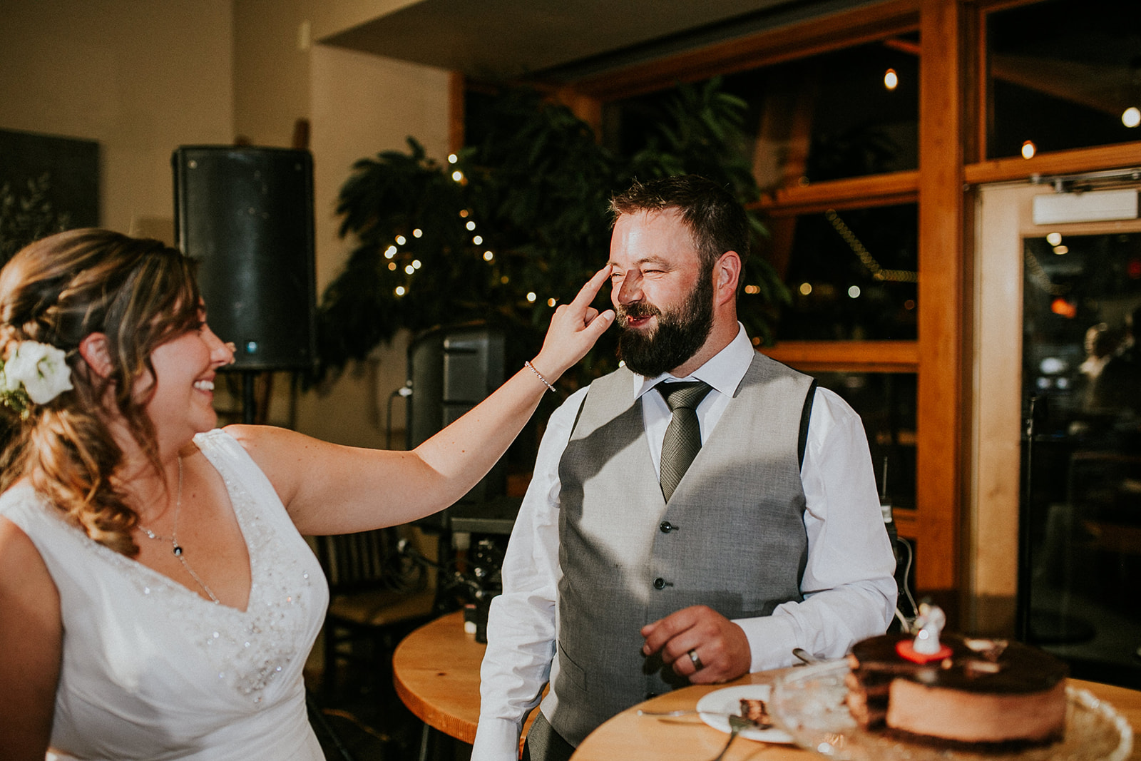 A bride playfully touches the groom’s face as they smile during their Canmore wedding reception. The groom stands beside a table with a chocolate cake, and the atmosphere is warm and festive.