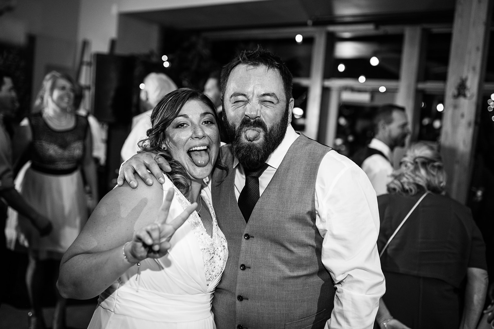 A woman in a white dress and a man in a vest and tie smile and make funny faces at the camera during a lively Calgary elopement, with people socializing in the background.