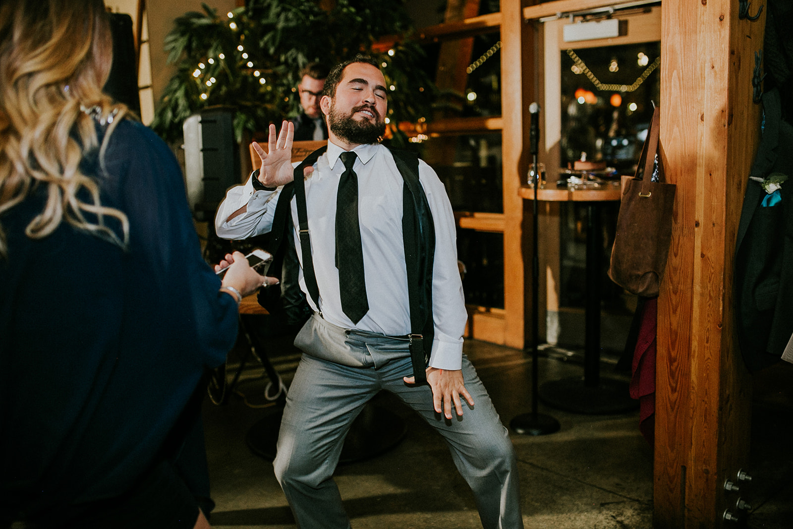 A man in a suit and tie dances enthusiastically indoors, eyes closed and one hand raised—captured by a talented photographer at a lively wedding. People, plants, and festive lights set the joyful scene in the background.