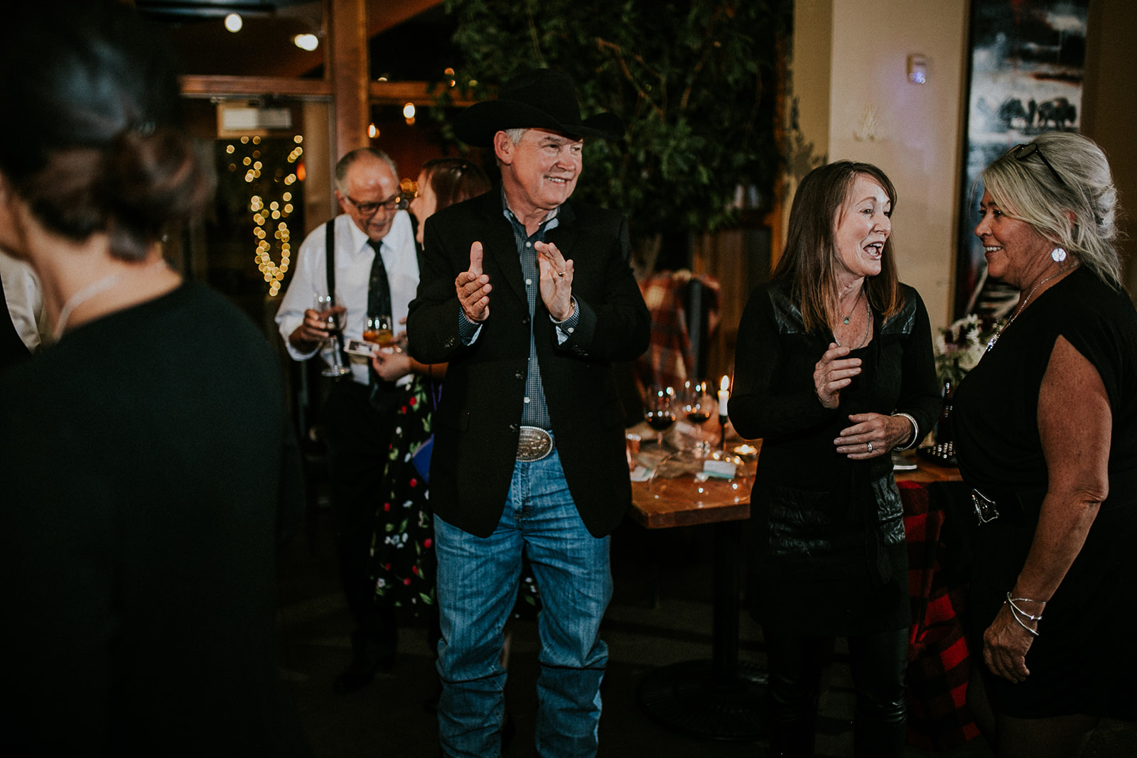 A man in a cowboy hat claps while two women laugh and talk at a lively indoor wedding. Other guests mingle in the background near decorated tables and string lights, as a photographer captures the joyful moments.