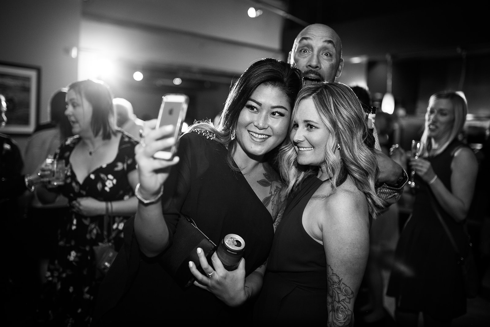 Three people pose for a selfie at a lively indoor event in Calgary. Two women smile at the camera while a man playfully photobombs them from behind, with others socializing in the background.