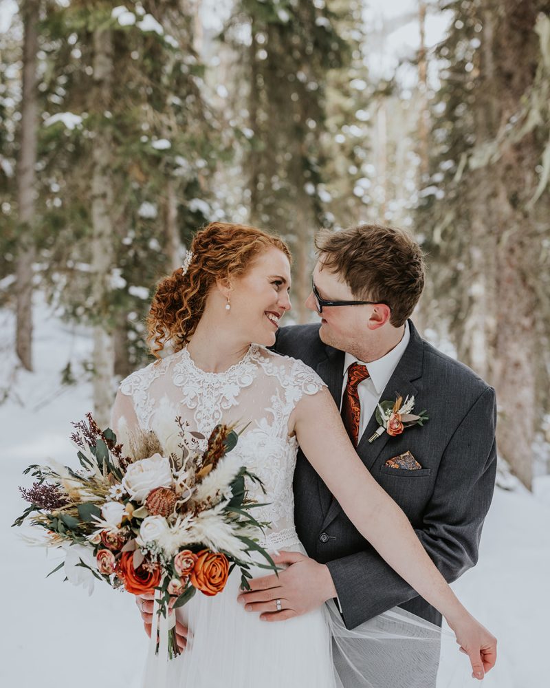 A bride in a lace dress holding a bouquet of orange and white flowers lovingly embraces her groom in a suit and tie. They share a joyful smile, standing in a snowy Banff forest surrounded by tall trees on their intimate elopement day.