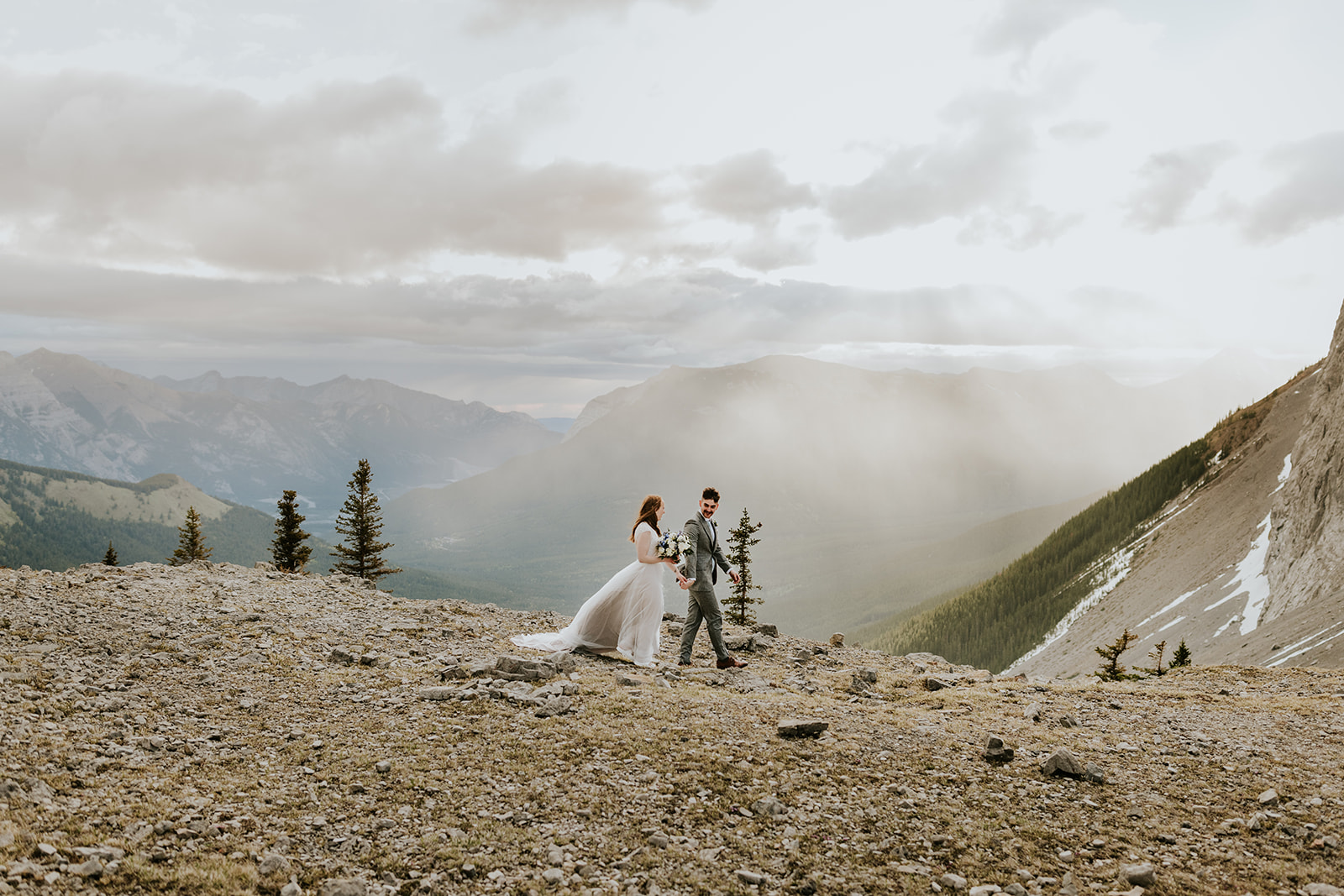 A bride and groom walk hand in hand on a rocky mountain landscape near Banff, surrounded by distant peaks and trees under a cloudy sky with sunlight streaming through—a breathtaking elopement moment.