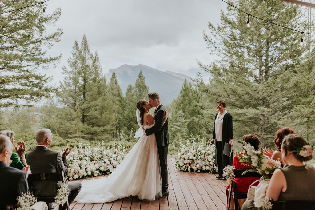 A bride and groom kiss at an outdoor wedding ceremony on a wooden deck surrounded by flowers and tall pine trees, with guests seated and clapping. A mountain is visible in the background under a cloudy sky.