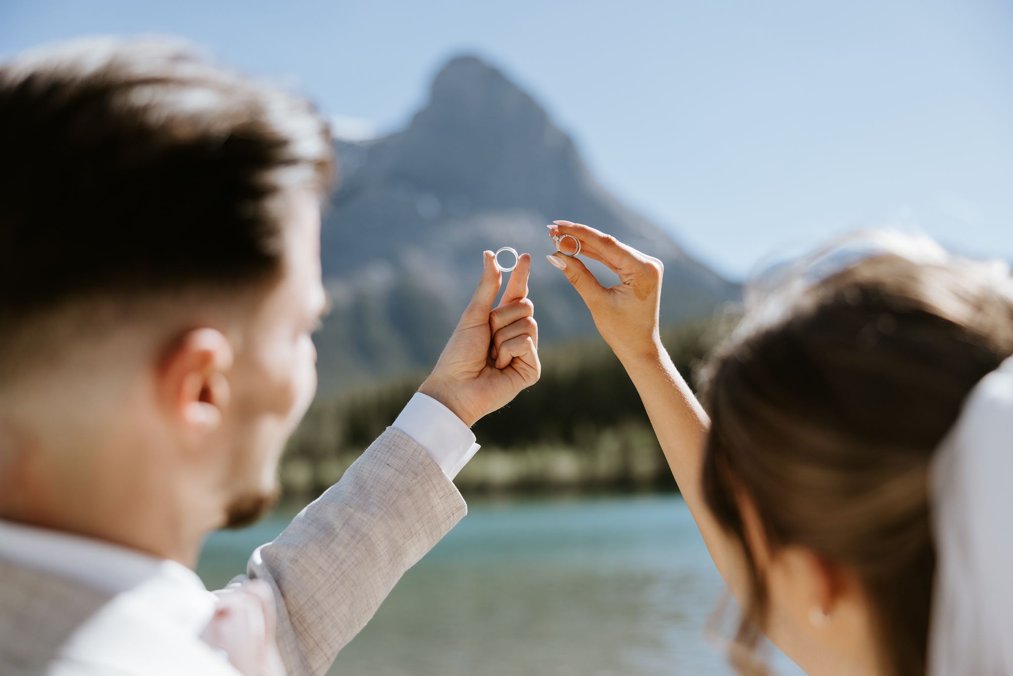 A couple stands by a lake, holding up wedding rings together with a mountain in the background, both slightly out of focus, emphasizing the rings against the scenic outdoor backdrop.