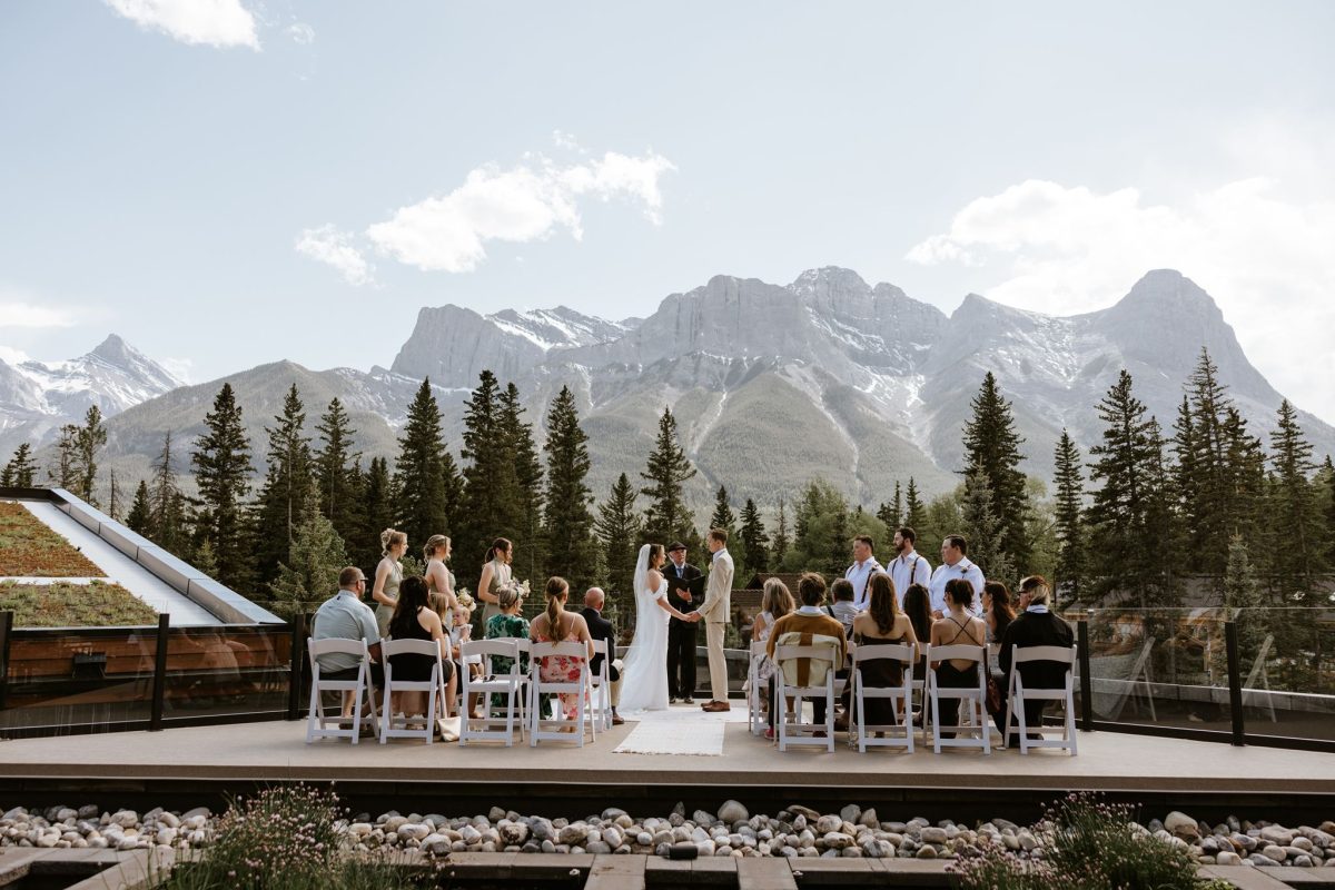 A wedding ceremony outdoors with guests seated, the couple and officiant standing, surrounded by pine trees and mountains under a clear sky.