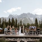 A wedding ceremony outdoors with guests seated, the couple and officiant standing, surrounded by pine trees and mountains under a clear sky.
