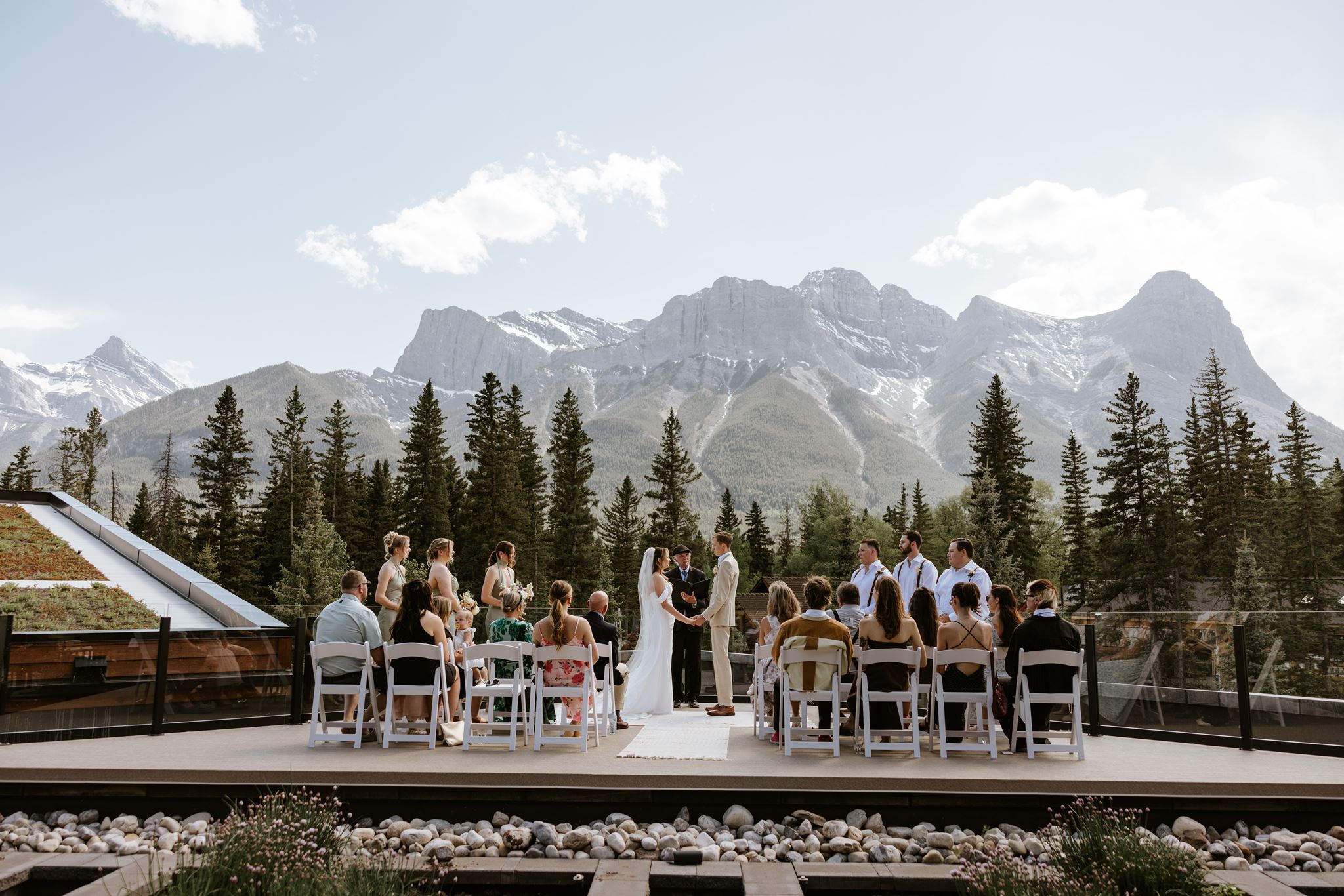 A wedding ceremony outdoors with guests seated, the couple and officiant standing, surrounded by pine trees and mountains under a clear sky.