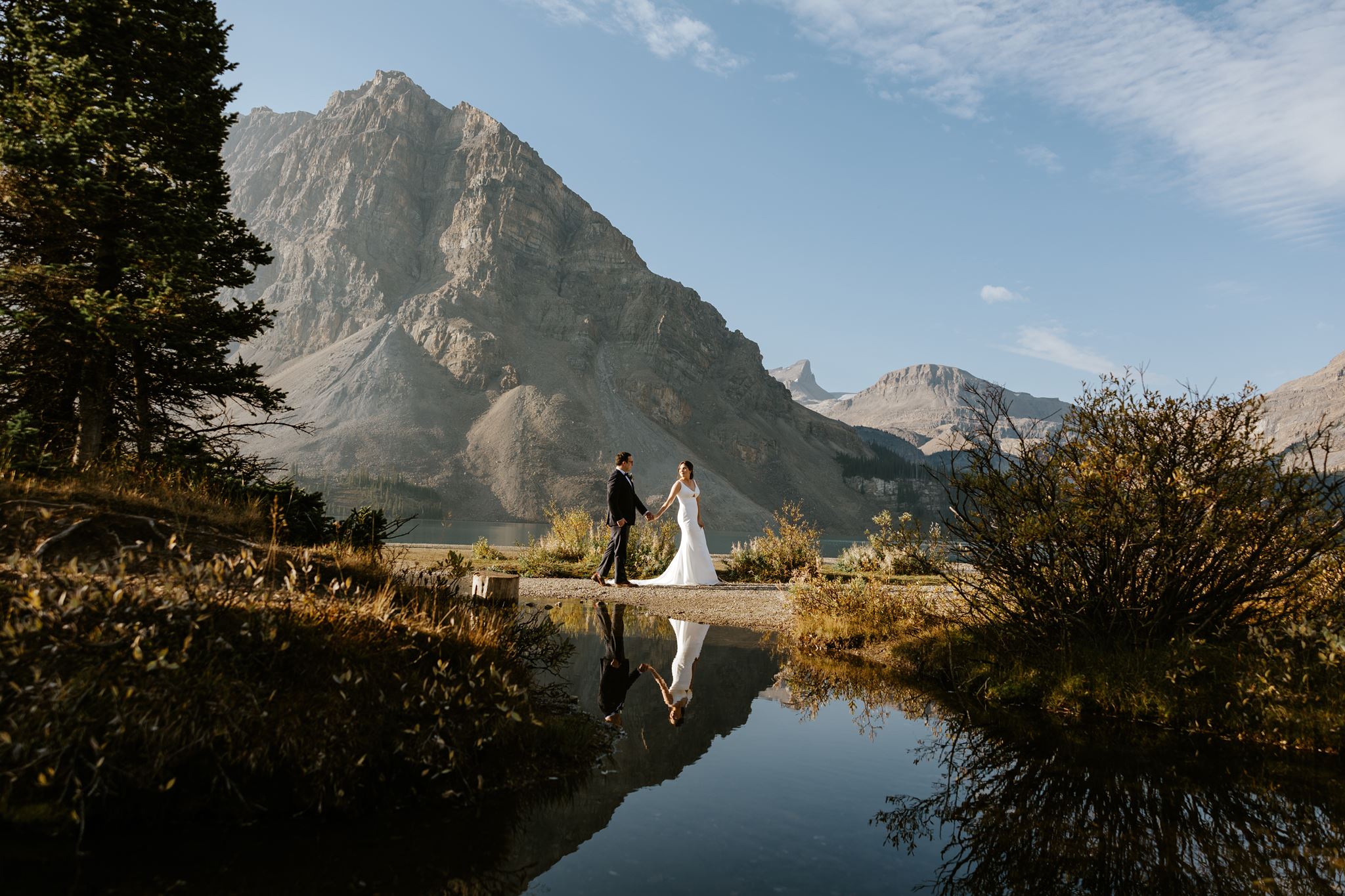 A bride and groom hold hands by a mountain lake, with their reflection in the water, surrounded by trees, shrubs, and dramatic mountain scenery under a partly cloudy sky.