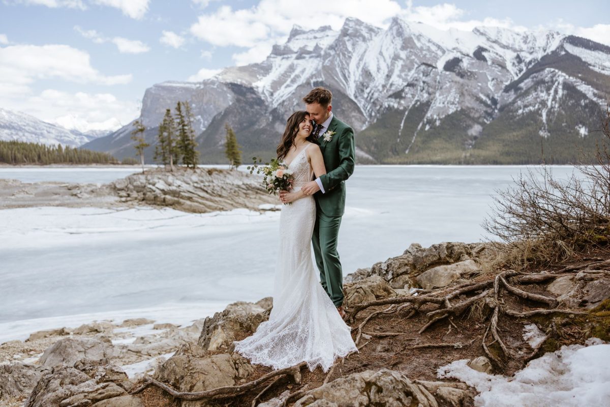 A bride and groom embrace on rocky ground by a frozen lake, with snow-capped mountains and trees in the background. The bride wears a white dress, and the groom wears a green suit.