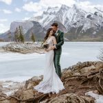 A bride and groom embrace on rocky ground by a frozen lake, with snow-capped mountains and trees in the background. The bride wears a white dress, and the groom wears a green suit.