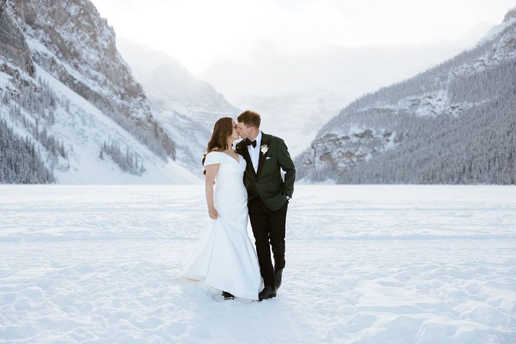 A bride and groom stand in the snow, dressed in wedding attire, embracing and smiling at each other with snowy mountains and trees in the background.