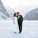 A bride and groom stand in the snow, dressed in wedding attire, embracing and smiling at each other with snowy mountains and trees in the background.
