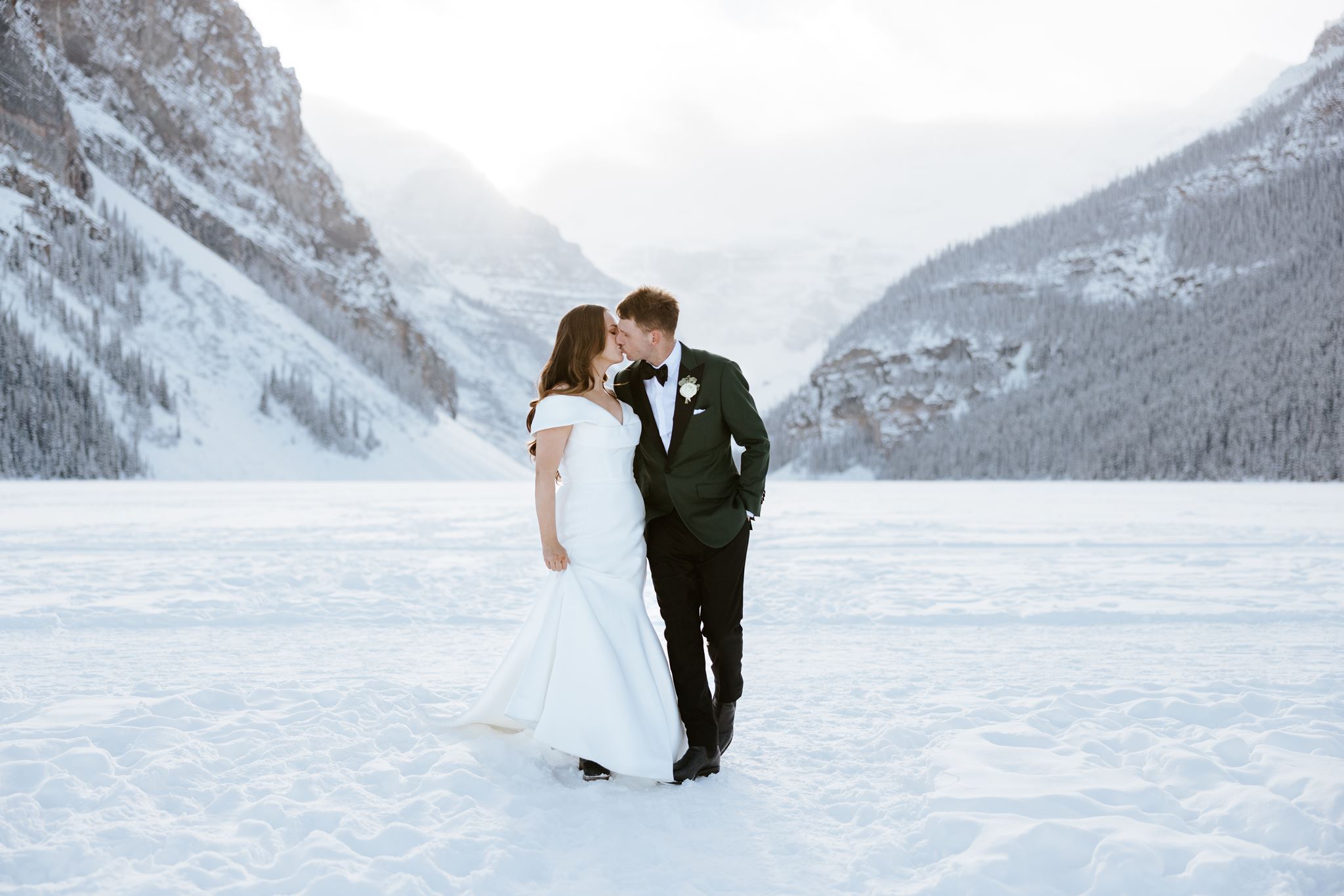 A bride and groom stand in the snow, dressed in wedding attire, embracing and smiling at each other with snowy mountains and trees in the background.