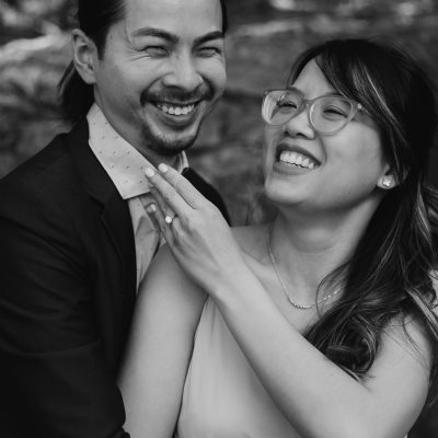 A couple smiles and laughs together outdoors in Calgary. The woman, wearing glasses and a ring, gently rests her hand on the suited man's chest. Captured by an elopement photographer, both appear joyful and happy in the black-and-white photo.