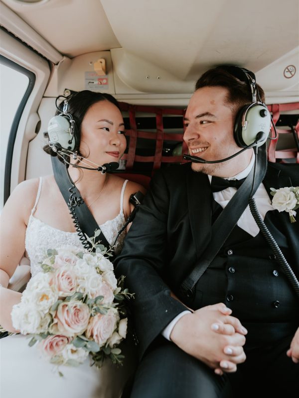 A bride and groom wearing headsets sit inside a helicopter over Banff, holding hands and smiling at each other. The bride, clutching a bouquet of pale pink and white roses, looks thrilled as their wedding photographer captures the joyful moment.