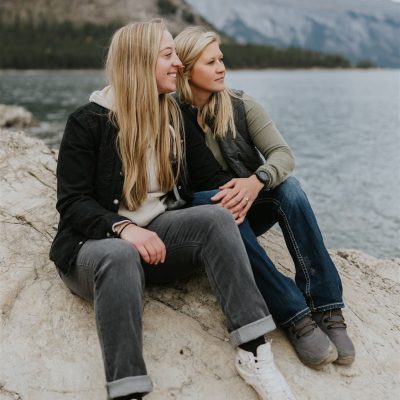 Two women with long blonde hair sit close together on a large rock by a lake, with mountains and trees in the background. Captured by a Calgary photographer, they appear relaxed, enjoying the natural scenery and peaceful atmosphere.