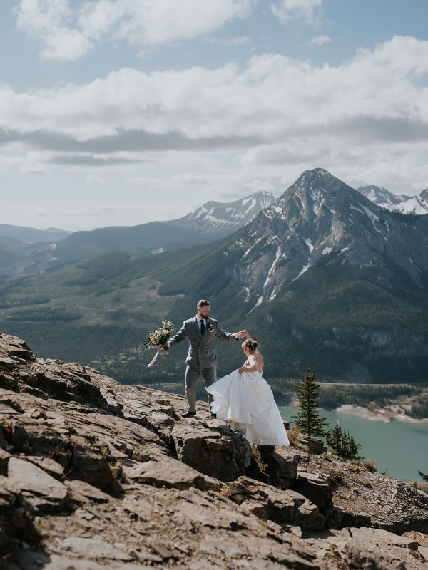 A couple in wedding attire stands on rocky mountain terrain, with the groom helping the bride up—an ideal elopement moment captured by a Calgary photographer, set against forested mountains, a lake, and a partly cloudy sky.
