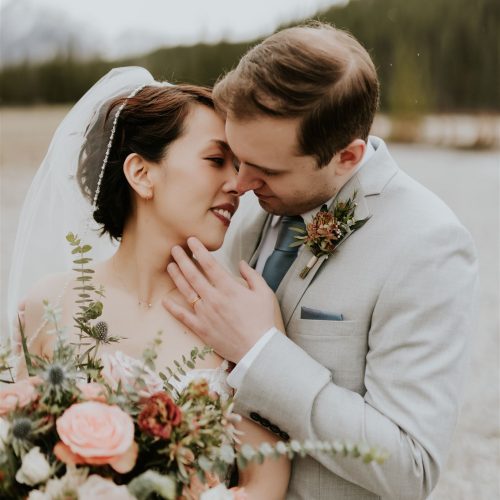 A bride and groom embrace outdoors in Canmore, smiling lovingly at each other. The bride holds a large bouquet of pink and white flowers, and both wear formal wedding attire with trees and mountains in the background.