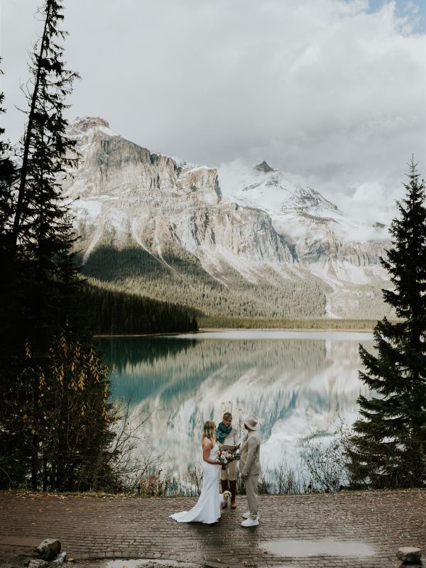 A couple stands on a stone path by a lake in Banff, exchanging elopement vows with a small child and officiant. Evergreen trees surround them as snow-capped mountains are reflected in the calm water under a partly cloudy sky, captured by their photographer.