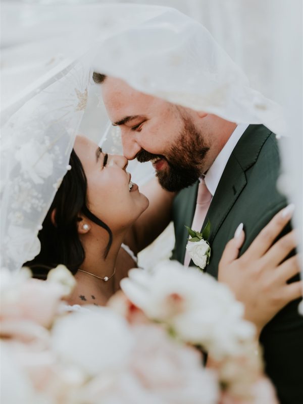 A joyful bride and groom smile and touch noses under a sheer white veil, surrounded by soft pastel flowers on their Calgary elopement wedding day.