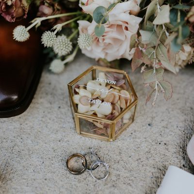 A hexagonal glass box filled with pale rose petals and labeled Devon & Taylor sits on a textured surface, surrounded by wedding rings, pearl jewelry, and a bouquet of soft-colored flowers—perfect for a romantic Banff or Canmore wedding.
