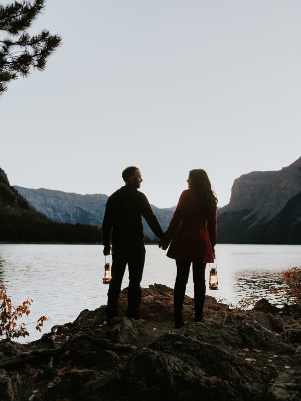 Silhouetted couple holding hands and lanterns stand on a rocky lakeshore at sunset in Canmore, facing calm water and mountains—captured perfectly by a wedding photographer under a clear sky.