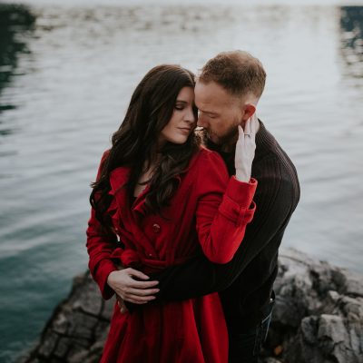 A couple embraces lovingly on a rocky Banff shoreline beside a calm lake. The woman, in a red coat, faces the camera while the man stands behind her, gently holding her—capturing an intimate elopement moment amidst breathtaking scenery.