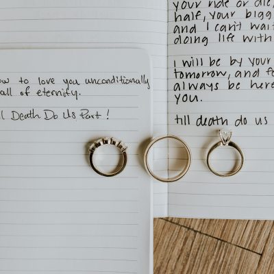 Three gold rings rest on open notebooks with handwritten wedding vows that include "till death do us part." White flowers lay beside them, captured beautifully by a Banff photographer to cherish these meaningful moments forever.