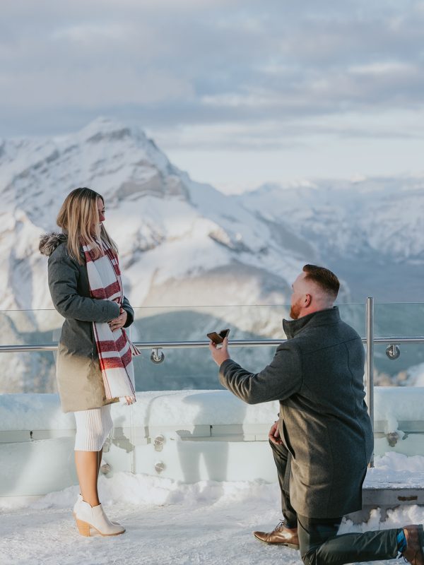 A man kneels on one knee in the snow, proposing with a ring to a smiling woman. Their elopement unfolds outdoors, with mountains and a cloudy sky creating the perfect backdrop for this romantic moment.