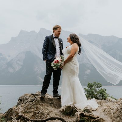 A bride and groom stand on a rocky outcrop by a lake near Canmore, facing each other. The bride’s veil flows in the wind as a wedding photographer captures the moment, with mountains rising under an overcast sky.