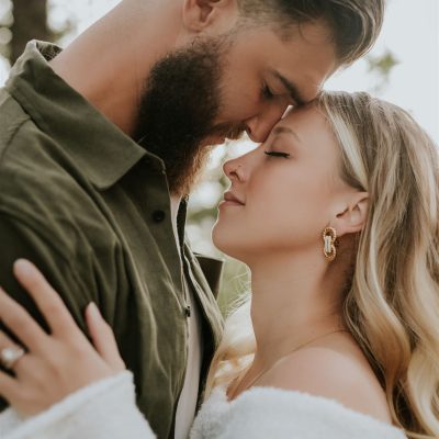 A man and woman embrace closely outdoors in Calgary, touching foreheads with eyes closed, sharing an intimate, tender moment. Captured by a photographer, he wears a green shirt and beard; she has long blonde hair and gold earrings.
