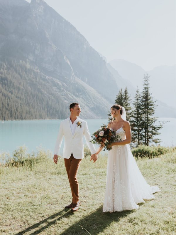 A bride in a white dress and a groom in a white jacket and brown pants hold hands, smiling at each other during their Canmore elopement, with a mountain lake and pine trees in the background.