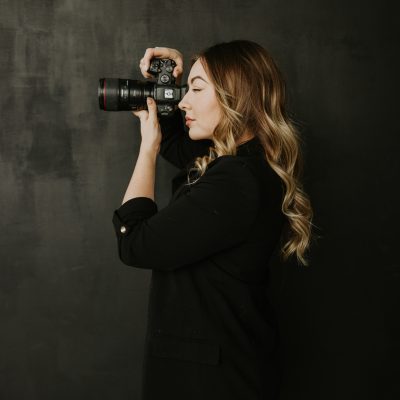A woman with long wavy hair, wearing a black blazer and grey pants, holds a camera up to her face and takes a photo against a dark, plain background—capturing the poised confidence of a Banff wedding photographer.