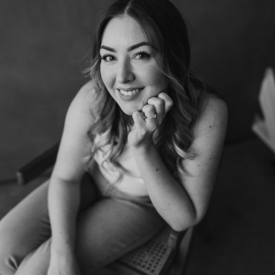 A woman with wavy hair sits on a chair, smiling at the camera with her chin resting on her hand. She wears a sleeveless top and jeans. The black-and-white photo captures a soft, natural setting, evoking the intimate feel of an elopement.