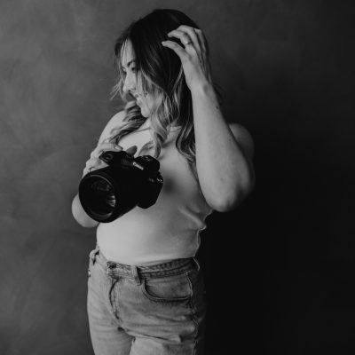 A woman in a sleeveless top and jeans holds a camera, looking down and adjusting her hair, standing against a plain dark background in a black and white photo—perfectly capturing the quiet anticipation before a Canmore elopement.