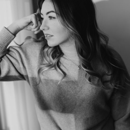 A young woman with wavy hair gazes thoughtfully out a window in Calgary, resting her elbow on the sill and her hand near her temple. She wears a light sweater, softly lit by natural light in this black-and-white photo.