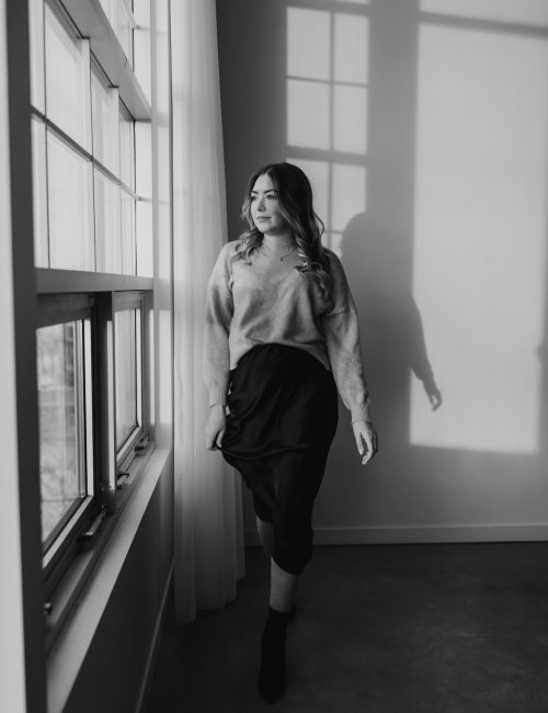 A woman in a sweater and midi skirt stands by large windows, looking outside. Soft light casts shadows on the wall behind her. Captured by a Canmore photographer, the black and white photo evokes a calm, contemplative mood.