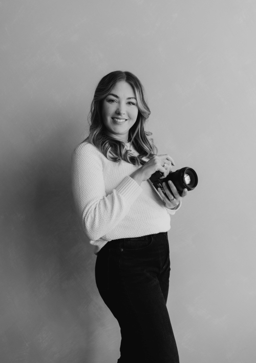 A woman with wavy hair, wearing a light sweater and dark pants, smiles while holding a camera—ready to capture an elopement. The background is plain and softly textured. The black-and-white photo adds timeless charm.