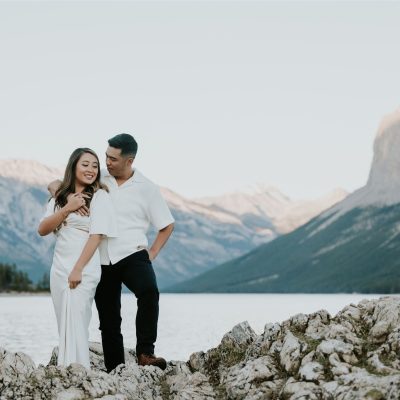 A couple stands on rocky terrain by a lake in Banff, smiling at each other. Snow-capped mountains and a forest provide the perfect backdrop for their wedding photographer under the clear sky.