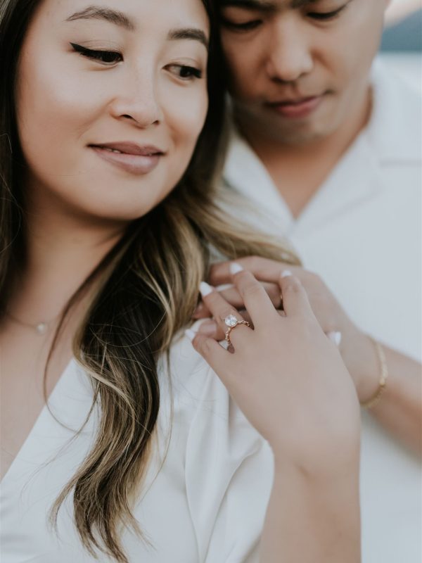 A close-up of a couple in a soft, intimate setting near Canmore. The woman, smiling softly, shows off a diamond engagement ring as the man lovingly embraces her from behind; both wear white clothing, capturing the romance of a wedding moment.