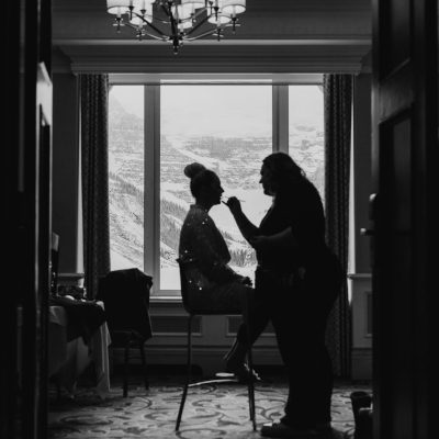 Black and white photo of a woman having her makeup done in a room with large windows, overlooking Calgary's snowy mountain landscape—an intimate elopement moment framed by an open doorway and a chandelier above.