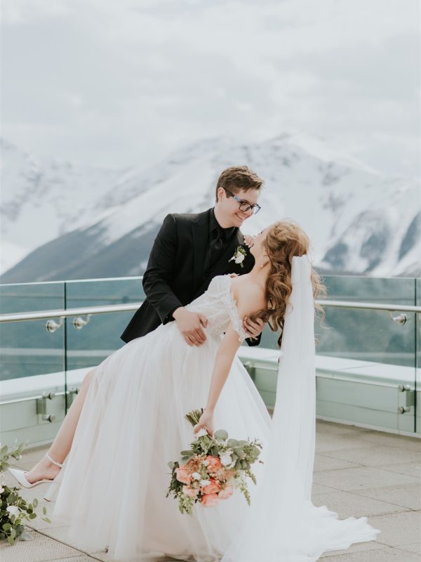 A groom in a black suit dips his bride in a white wedding dress and veil on a balcony, with snow-capped Banff mountains in the background. The bride holds a bouquet of flowers and they are smiling at each other on their wedding day.