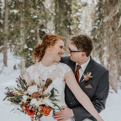 A bride in a lace dress holding a bouquet of orange and white flowers lovingly embraces her groom in a suit and tie. They share a joyful smile, standing in a snowy Banff forest surrounded by tall trees on their intimate elopement day.