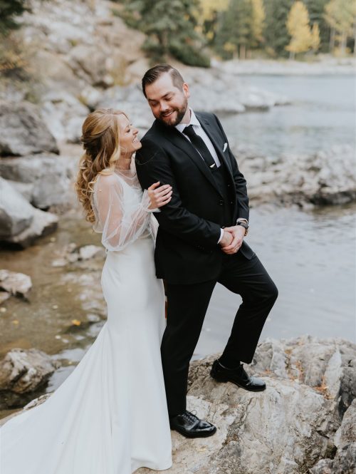A bride in a white dress and a groom in a black suit stand on rocks by a lakeshore, smiling and laughing together with trees and water in the background—captured by a Calgary photographer during their intimate elopement.