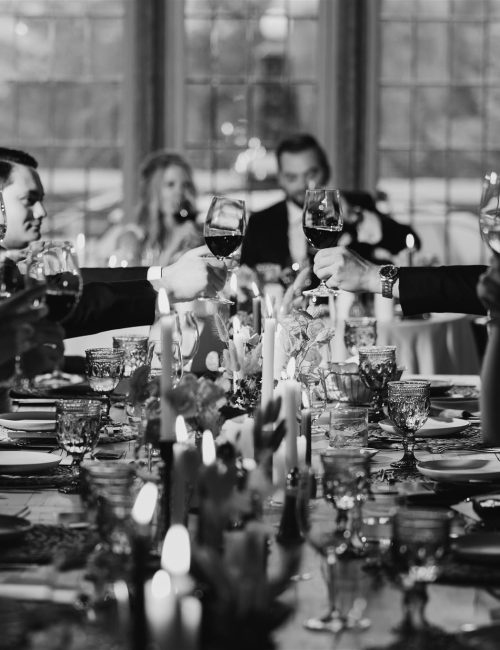 A group of people dressed formally sit around a long, elegant dinner table in Banff, raising their wine glasses for a toast. Captured by a skilled photographer, the black and white scene features candles and decorative settings.